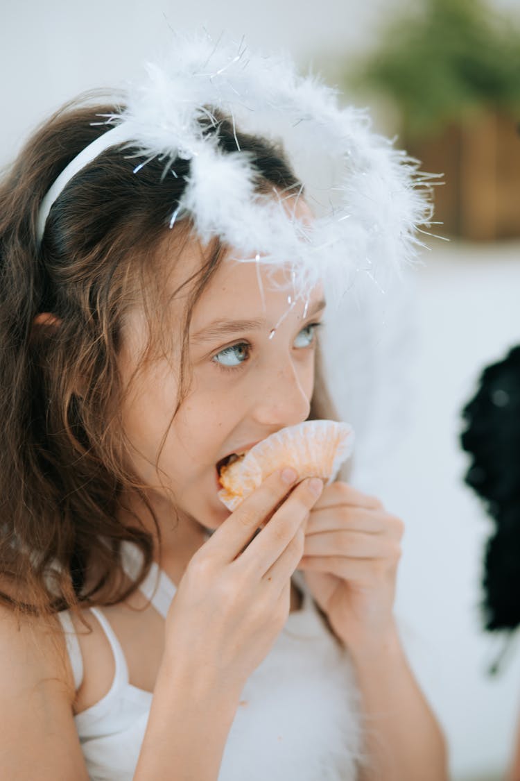 Young Brunette Girl In A Costume Of An Angel Eating A Cupcake And Looking Up