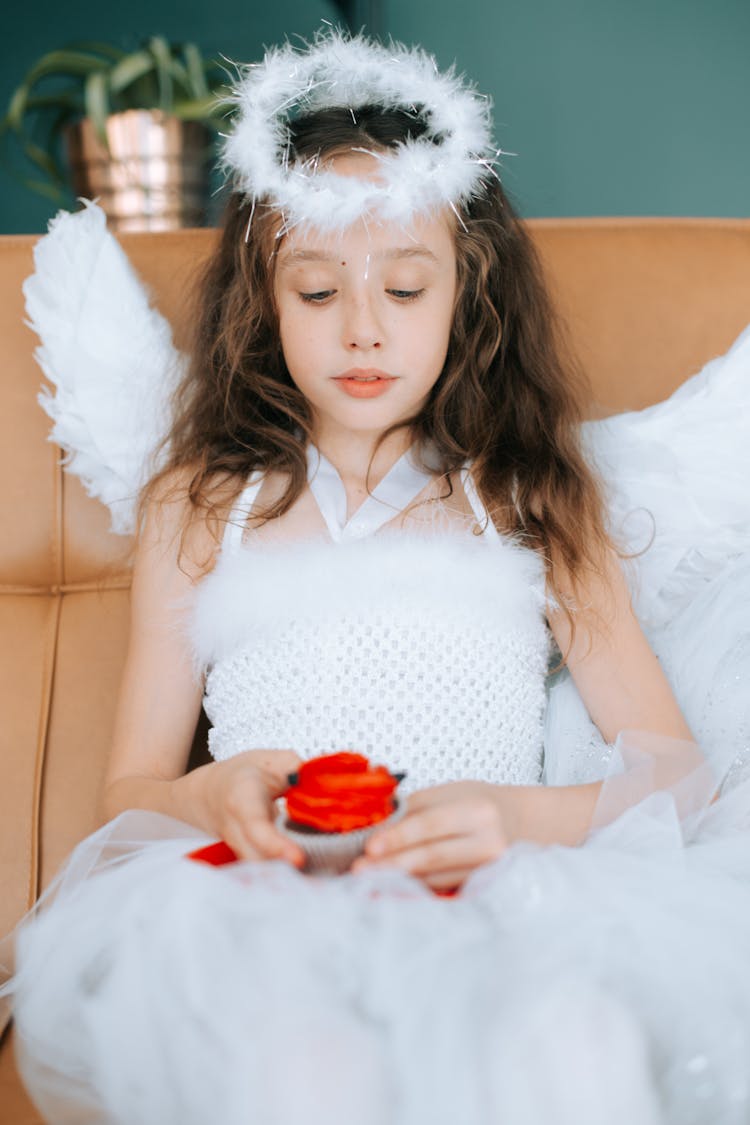 Young Brunette Girl In A Costume Of An Angel Holding A Cupcake