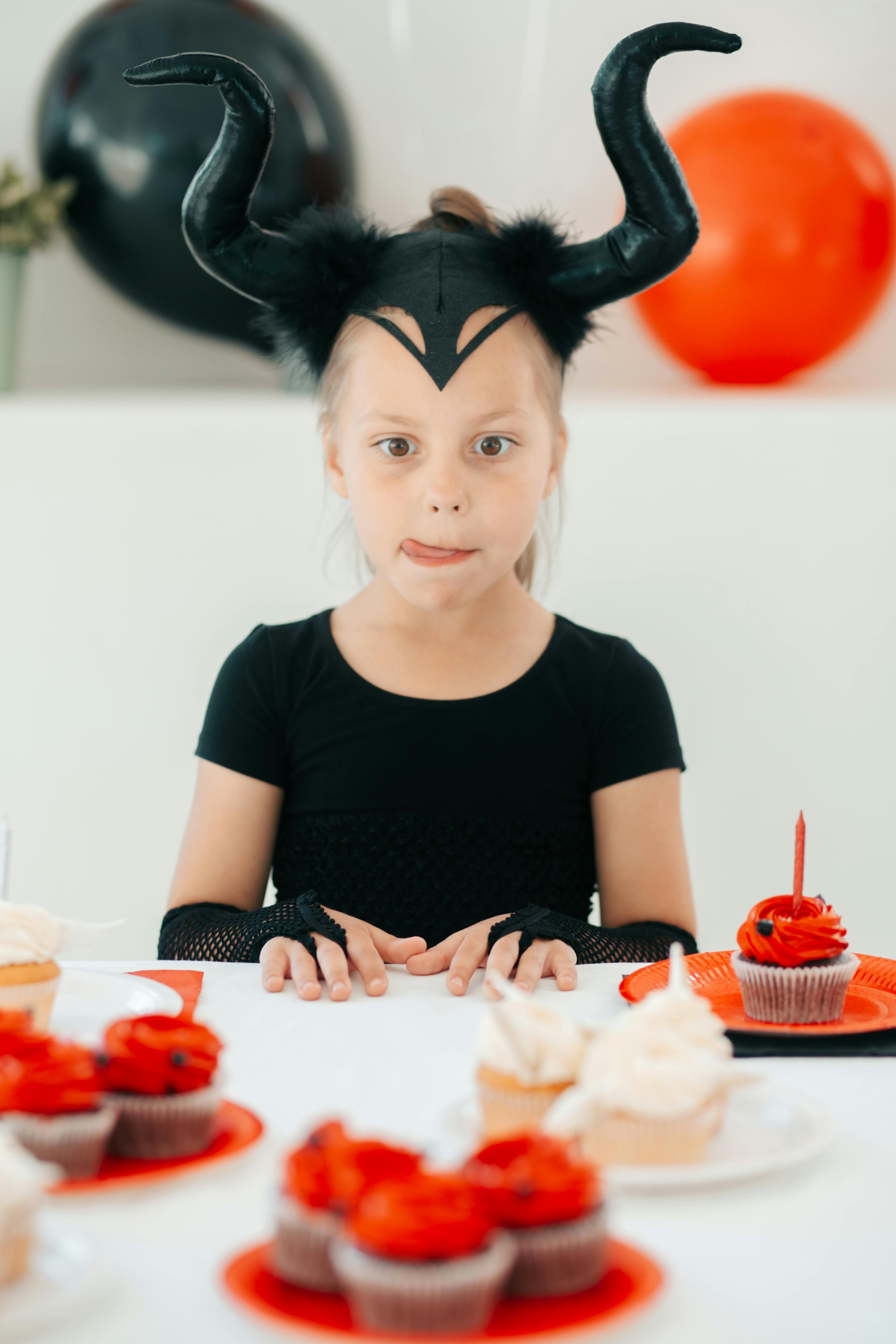 little girl in a costume with devil horns sitting at a table with sweets