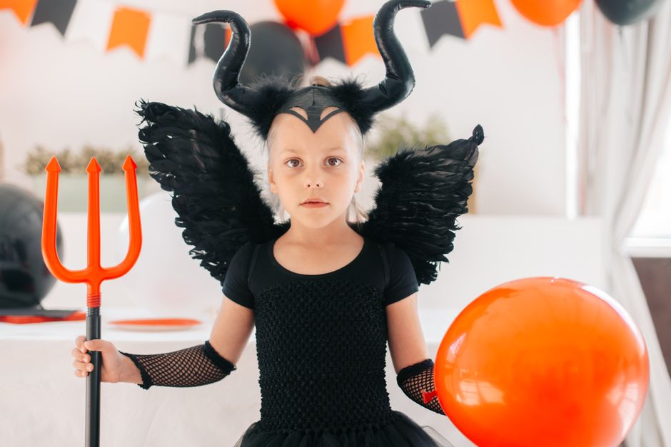 Cute child dressed as a devil with horns and wings, holding a trident and balloon indoors.