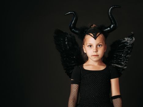 A young girl dressed in a black devil costume with wings and horns, on a dark background.