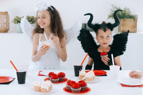 Free Kids dressed as angel and devil enjoy cupcakes at a Halloween party. Stock Photo