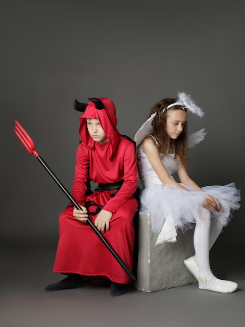 A Young Boy and Girl Wearing a Devil and Angel Costumes while Sitting on the Chair Back to Back