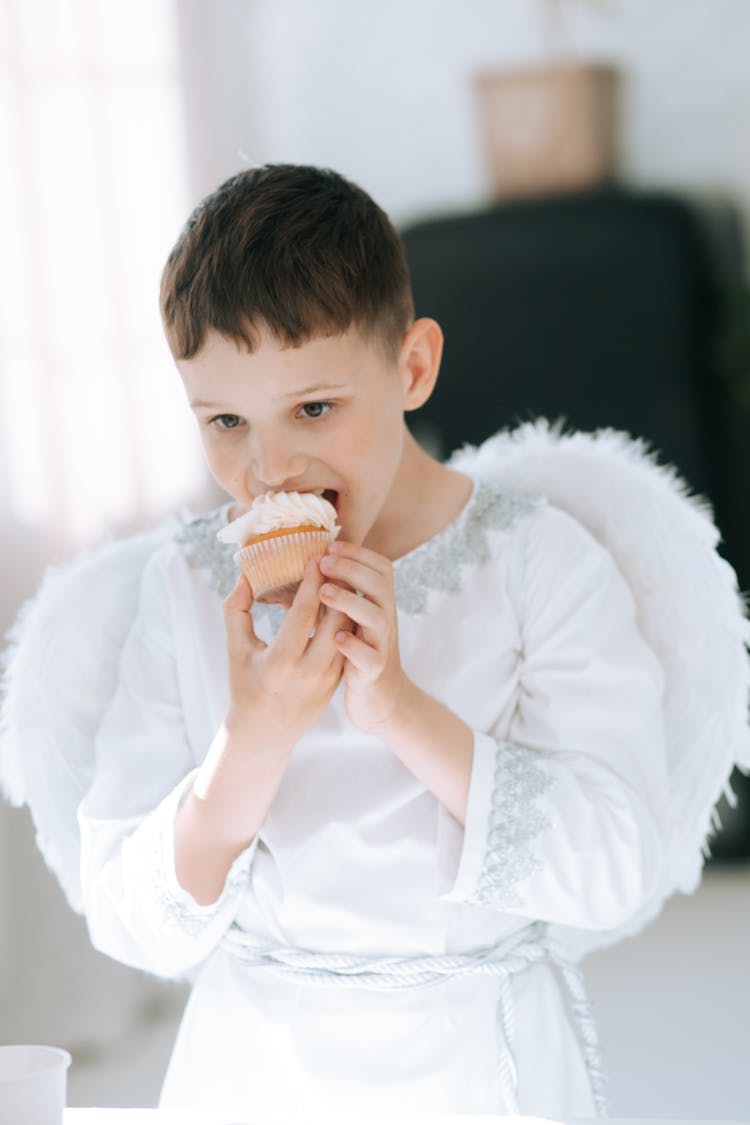 A Boy In Angel Costume Eating A Cupcake