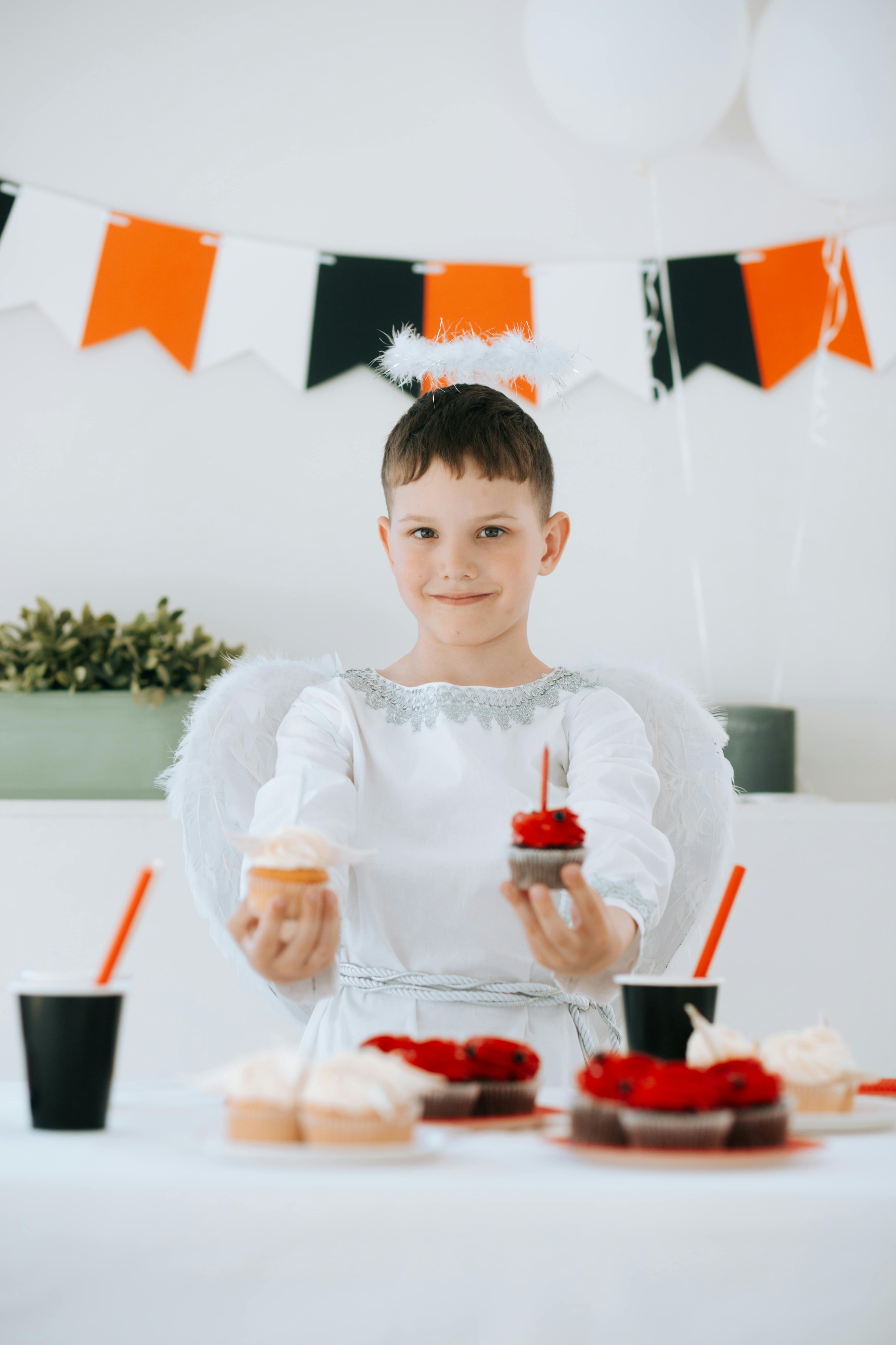 Young boy in angel costume holding cupcakes during a festive Halloween celebration.
