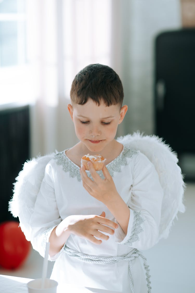 A Boy In Angel Costume Eating A Cupcake