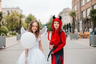 Delighted girl and boy in angel and devil costumes standing together in street on blurred background and looking at camera