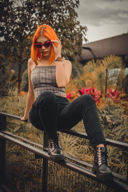 Free Stylish woman with orange hair and sunglasses posing on metal railing outdoors. Stock Photo