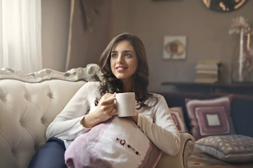Free Woman Wearing White Top Drinking Beverage from White Ceramic Mug While Lying on Sofa Inside Well Lit Room Stock Photo