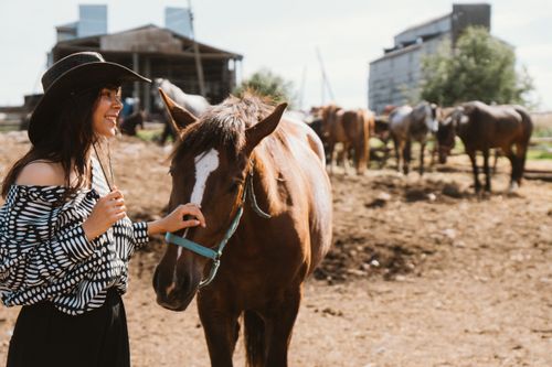 Free A woman in a stylish outfit smiles as she pets a horse on a sunny farm. Stock Photo