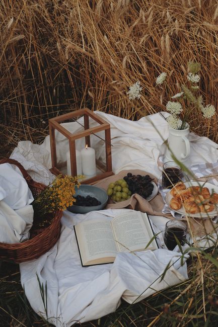 Cozy picnic setup with books, grapes, croissants, and decor in a golden wheat field.