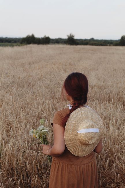 Woman with straw hat and flowers standing in a wheat field, back view.