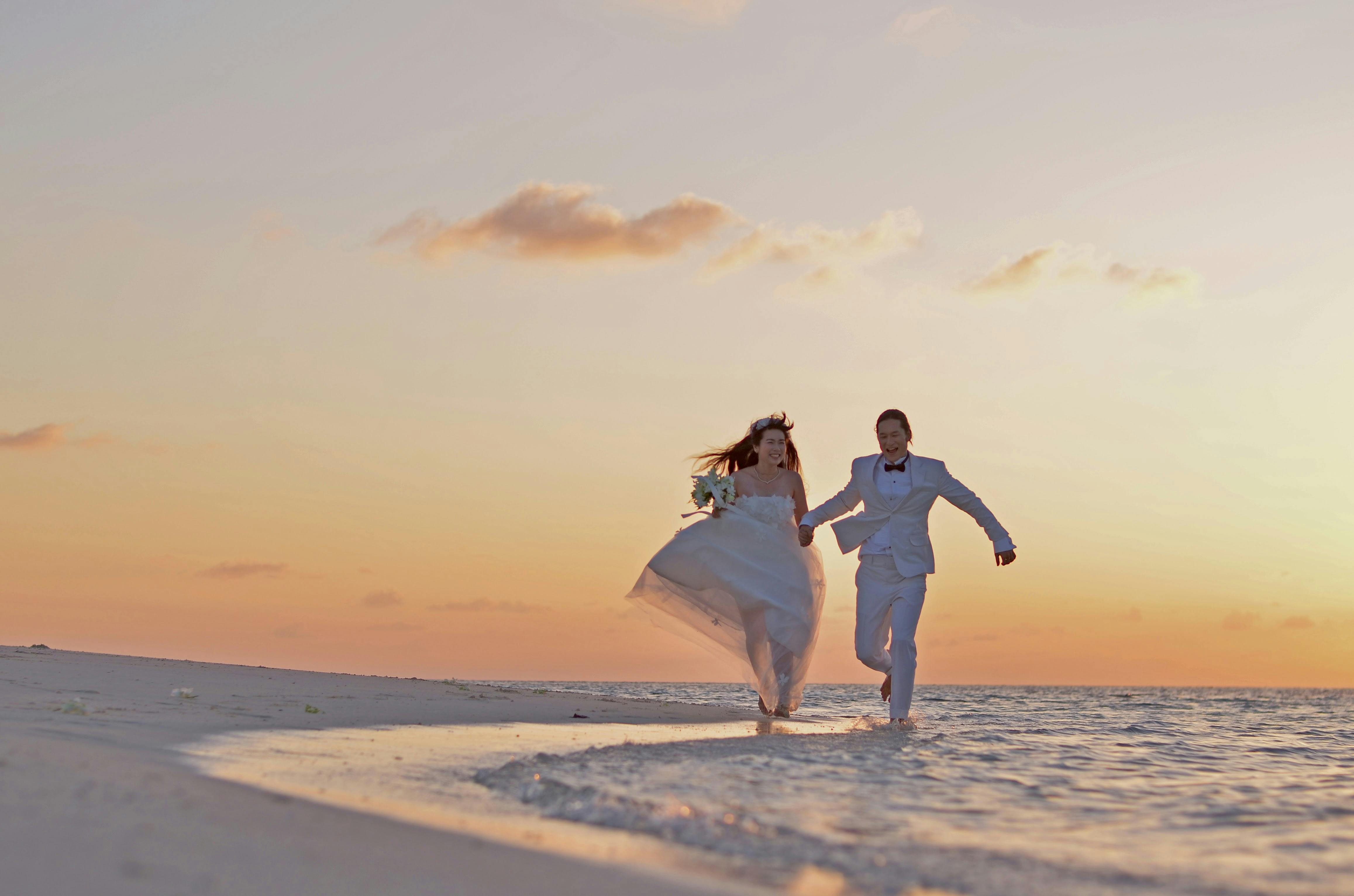 Newlywed couple joyfully running on a sandy beach during a beautiful sunset.
