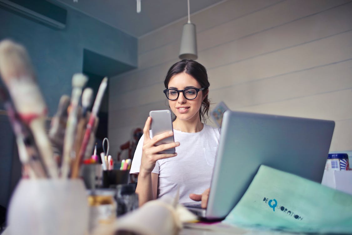 Free Woman in White T-shirt Holding Smartphone in Front of Laptop Stock Photo