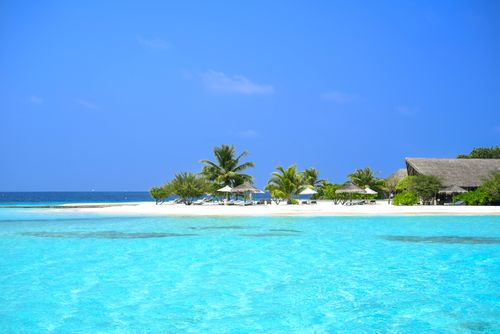 Free Stunning tropical beach with clear blue water, palm trees, and resort huts, perfect for a relaxing holiday. Stock Photo