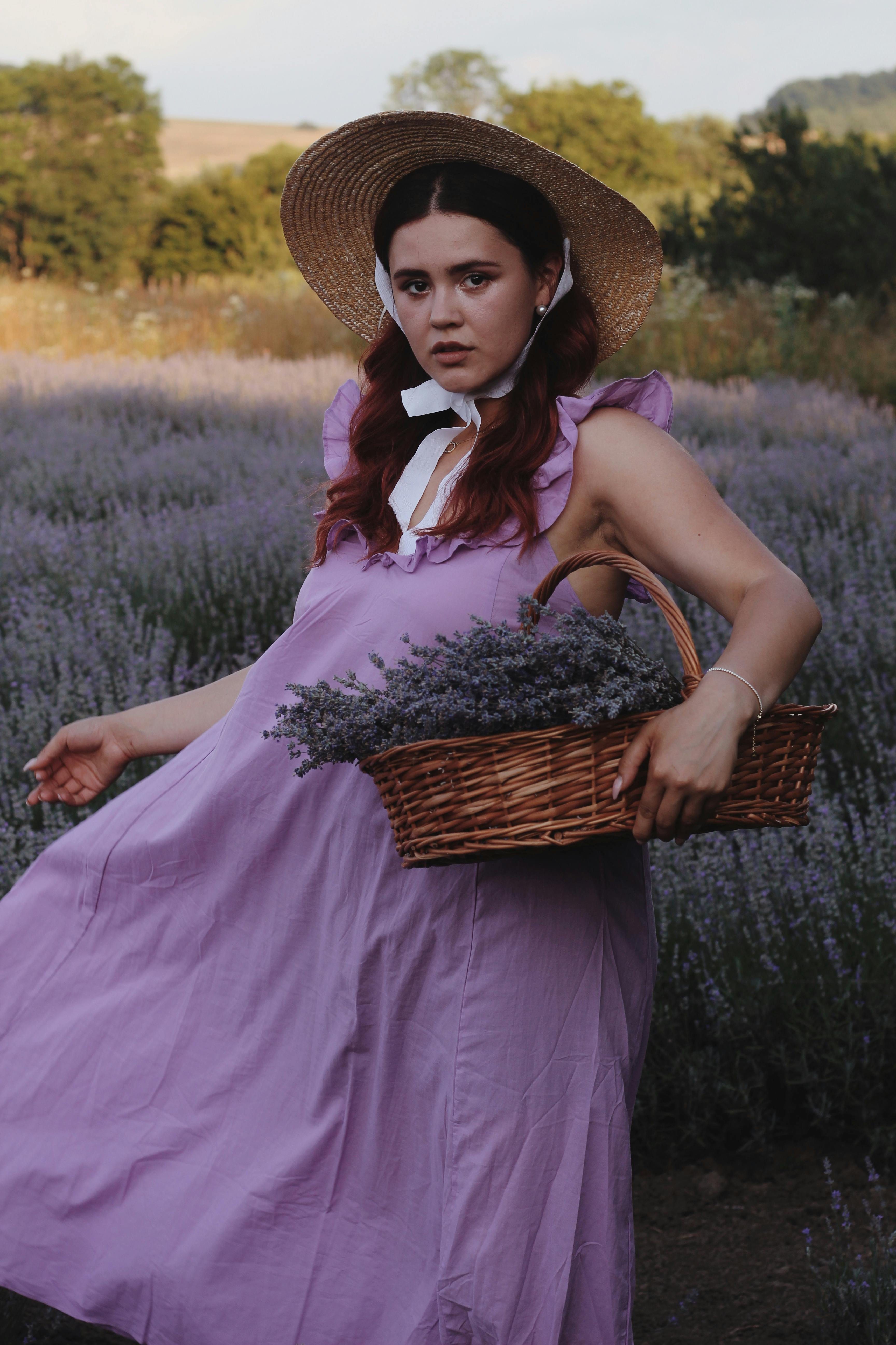 Elegant woman in purple dress and sun hat holding lavender in a wicker basket outdoors.