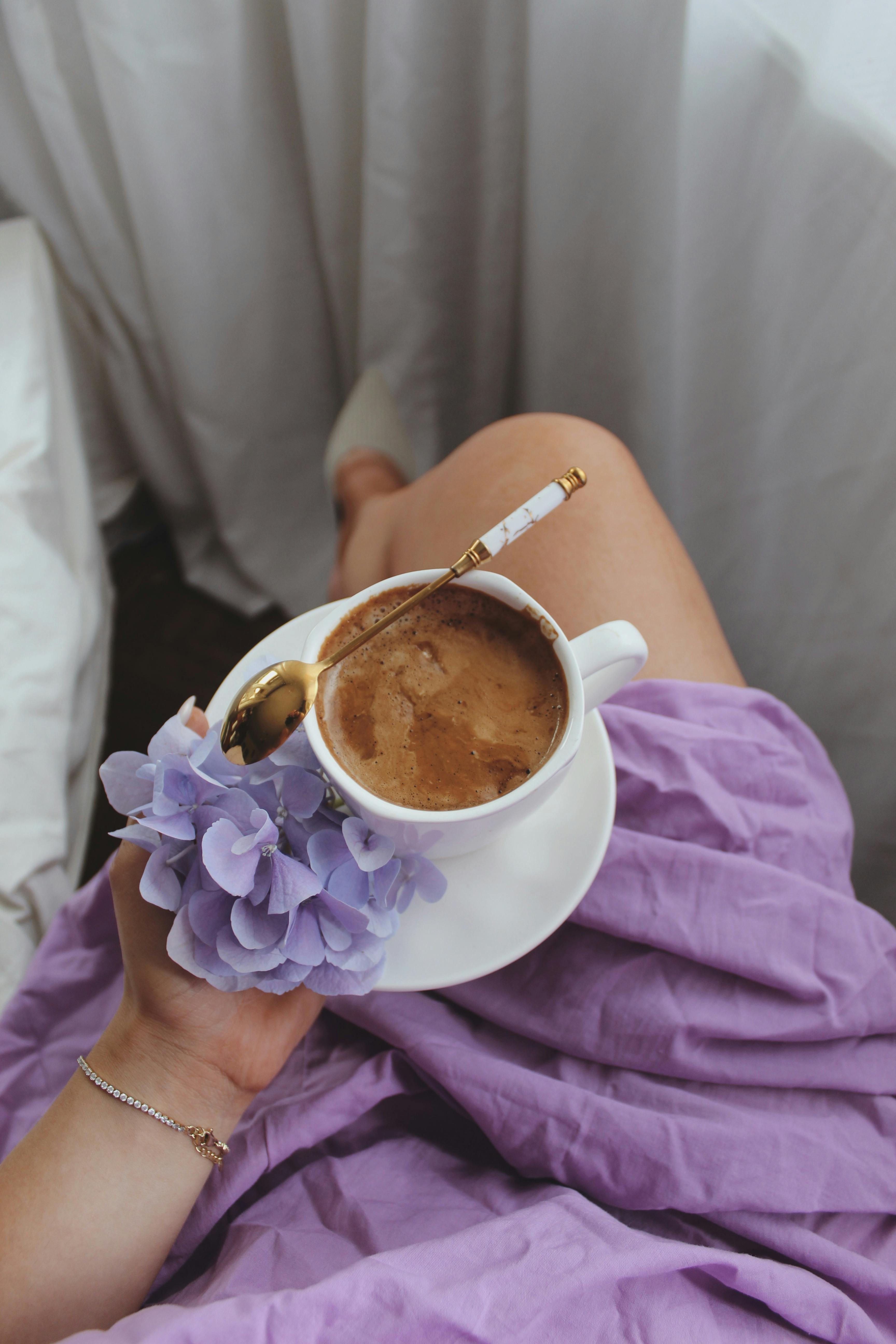 A cozy morning scene featuring a cup of coffee, violet flowers, and a woman's hand draped in purple fabric.