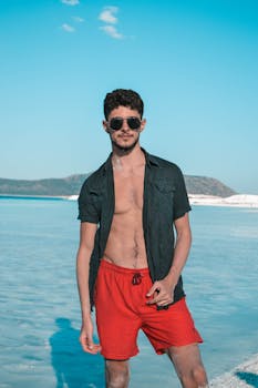 A young man in red shorts and sunglasses posing by the beach, showcasing summer vibes.