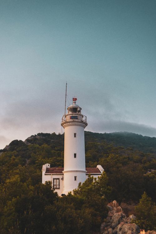 Free A serene lighthouse stands tall amidst lush hills under a clear blue sky with low clouds. Stock Photo