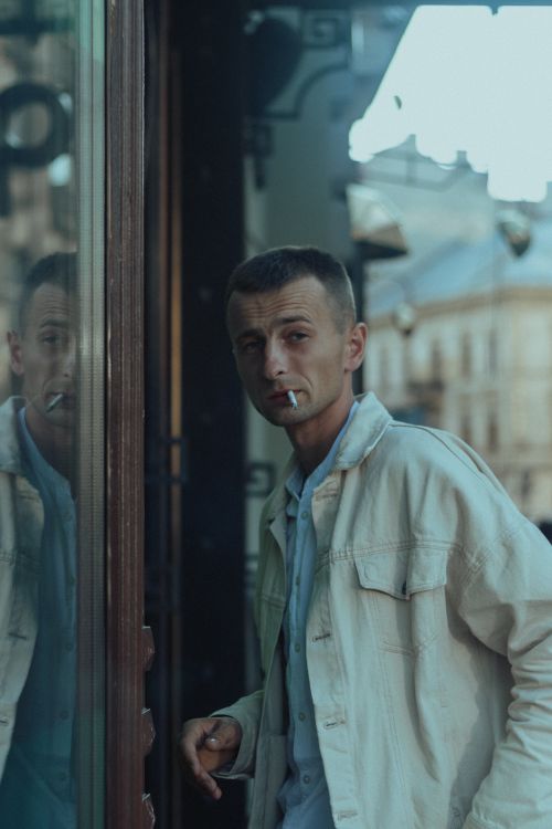 Free A man in a white jacket smokes while leaning against a glass wall on the street. Stock Photo