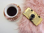 粉红色的皮毛表面上平放着一款时尚咖啡杯和一部金色相机主题的智能手机