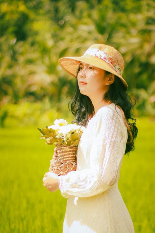 Free A woman in a white dress and sun hat holding flowers, standing in a lush green field. Stock Photo