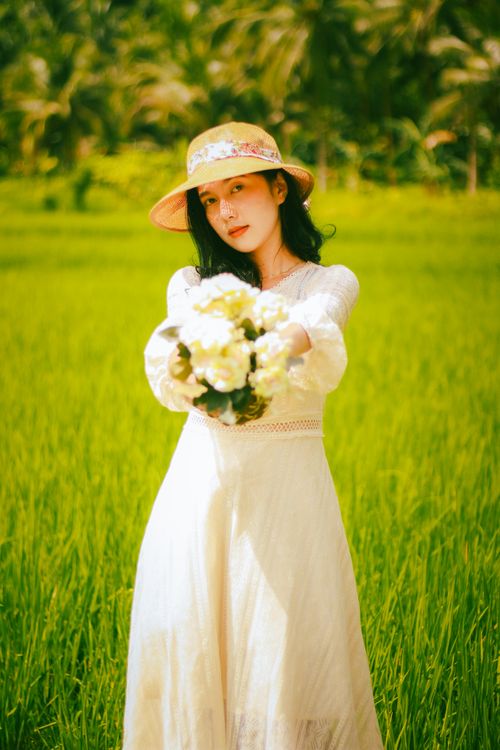 Free Woman in a white dress holds a bouquet in a lush green field on a sunny day. Stock Photo