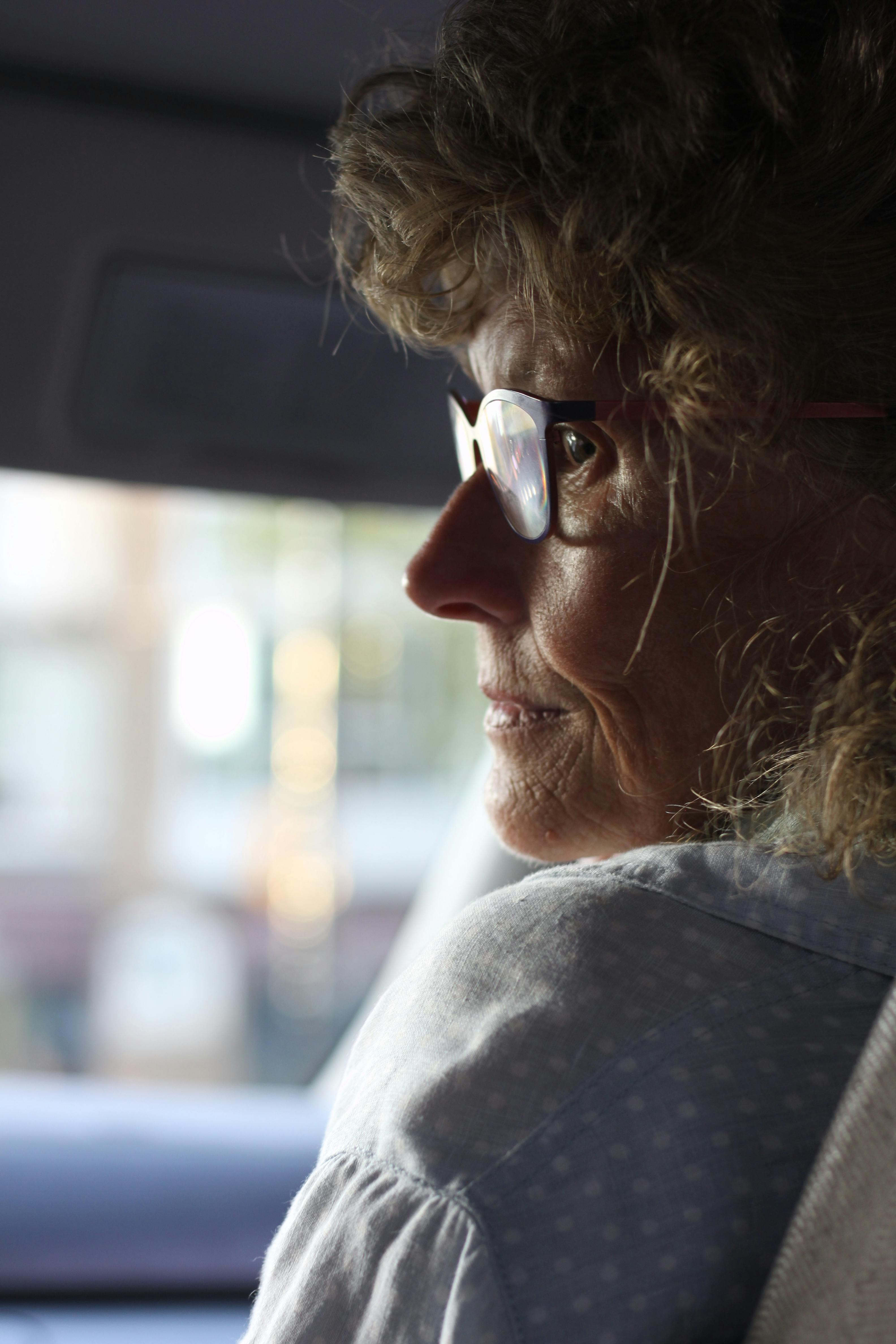 Close-up profile portrait of an elderly woman with eyeglasses inside a vehicle.