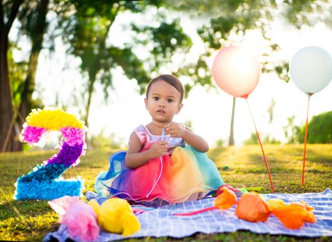 A toddler in a vibrant dress celebrates a birthday outdoors with balloons and decorations.