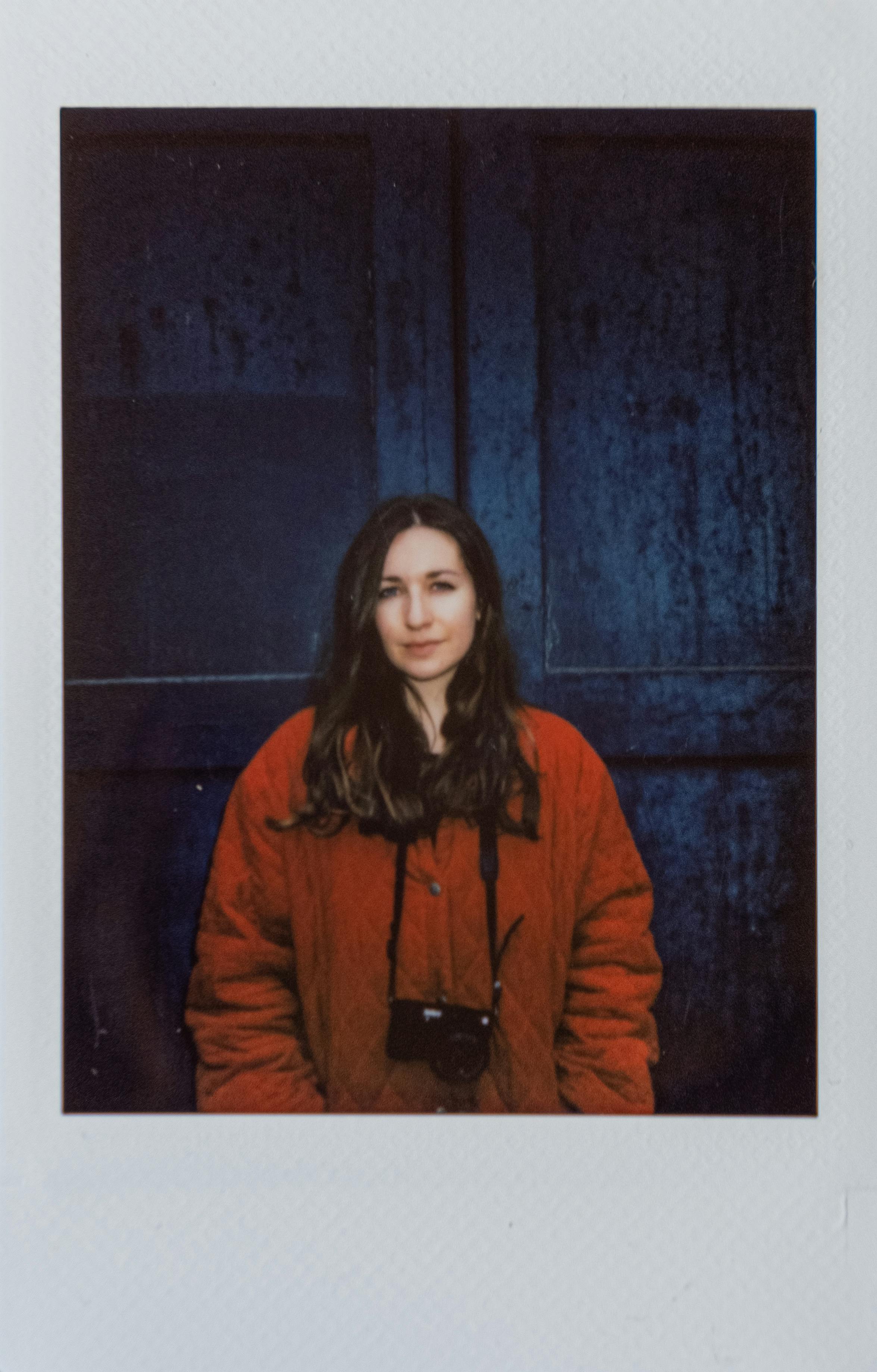 A woman in a red sweater stands against a dark backdrop holding a camera.