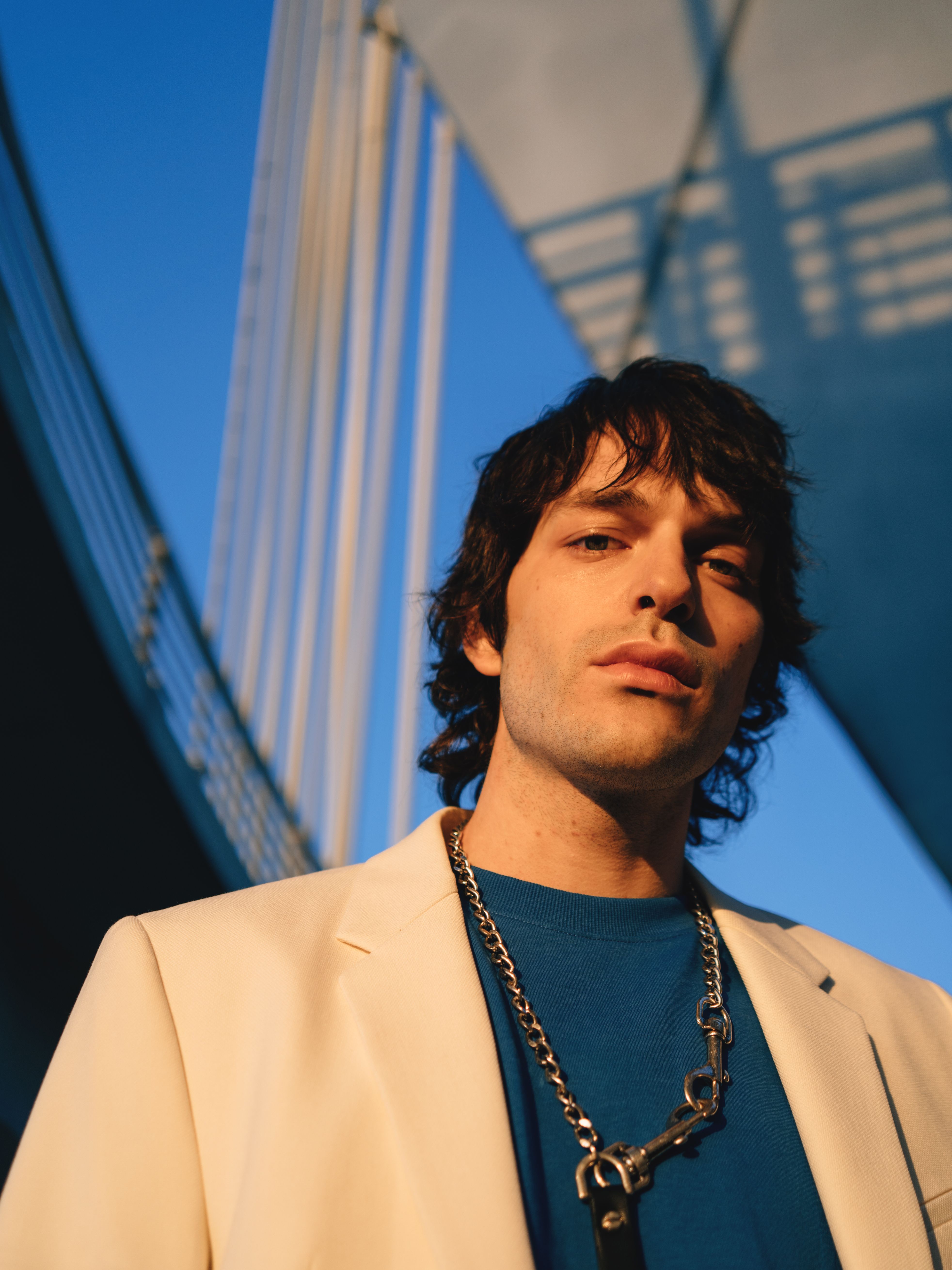 Portrait of a man in a white blazer and chain necklace with modern architecture in the background.