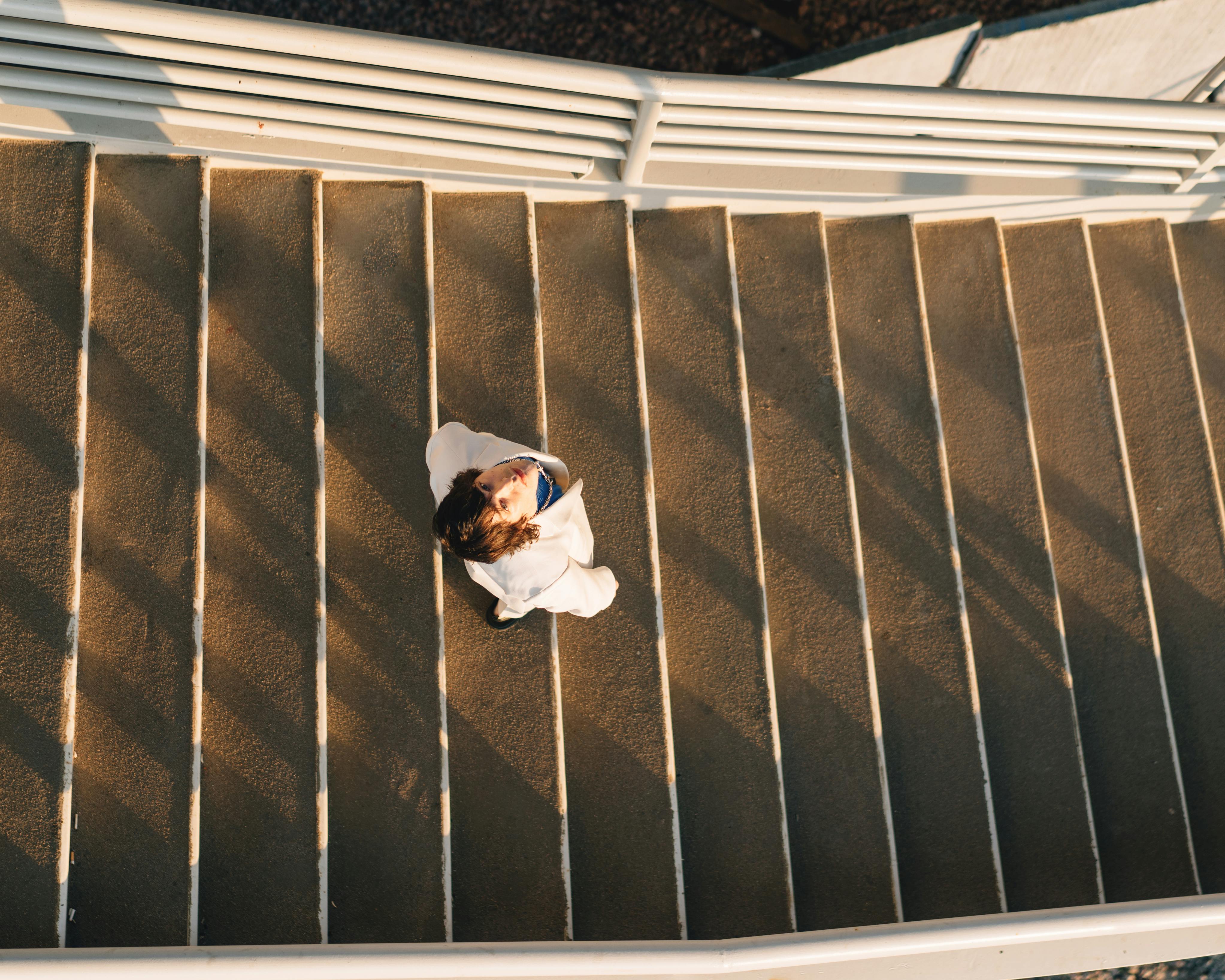 Top view of a man in a white jacket standing on stairs in an urban setting, casting shadows.