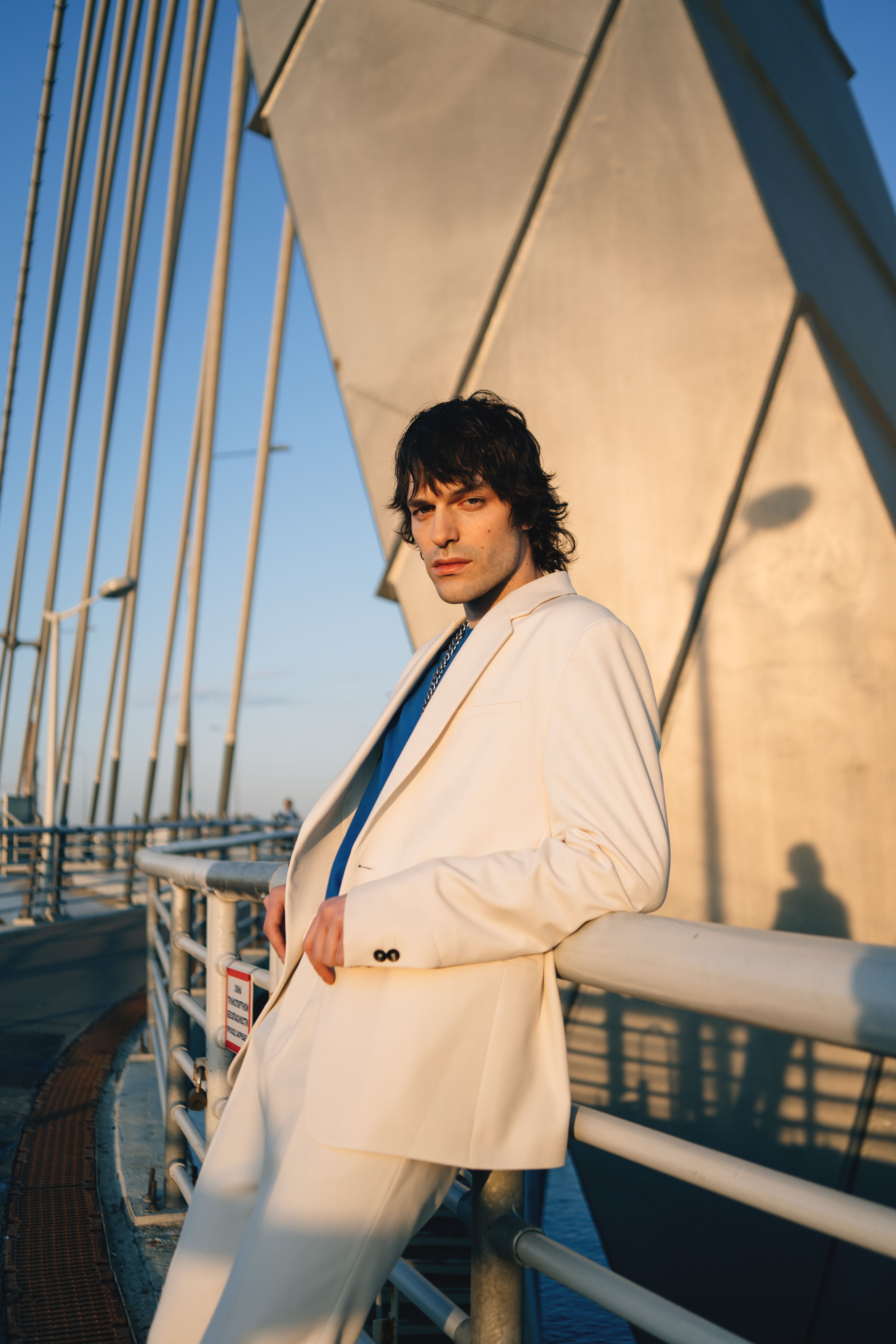 A man in a white suit jacket leans on a bridge railing during sunset, showcasing modern fashion.