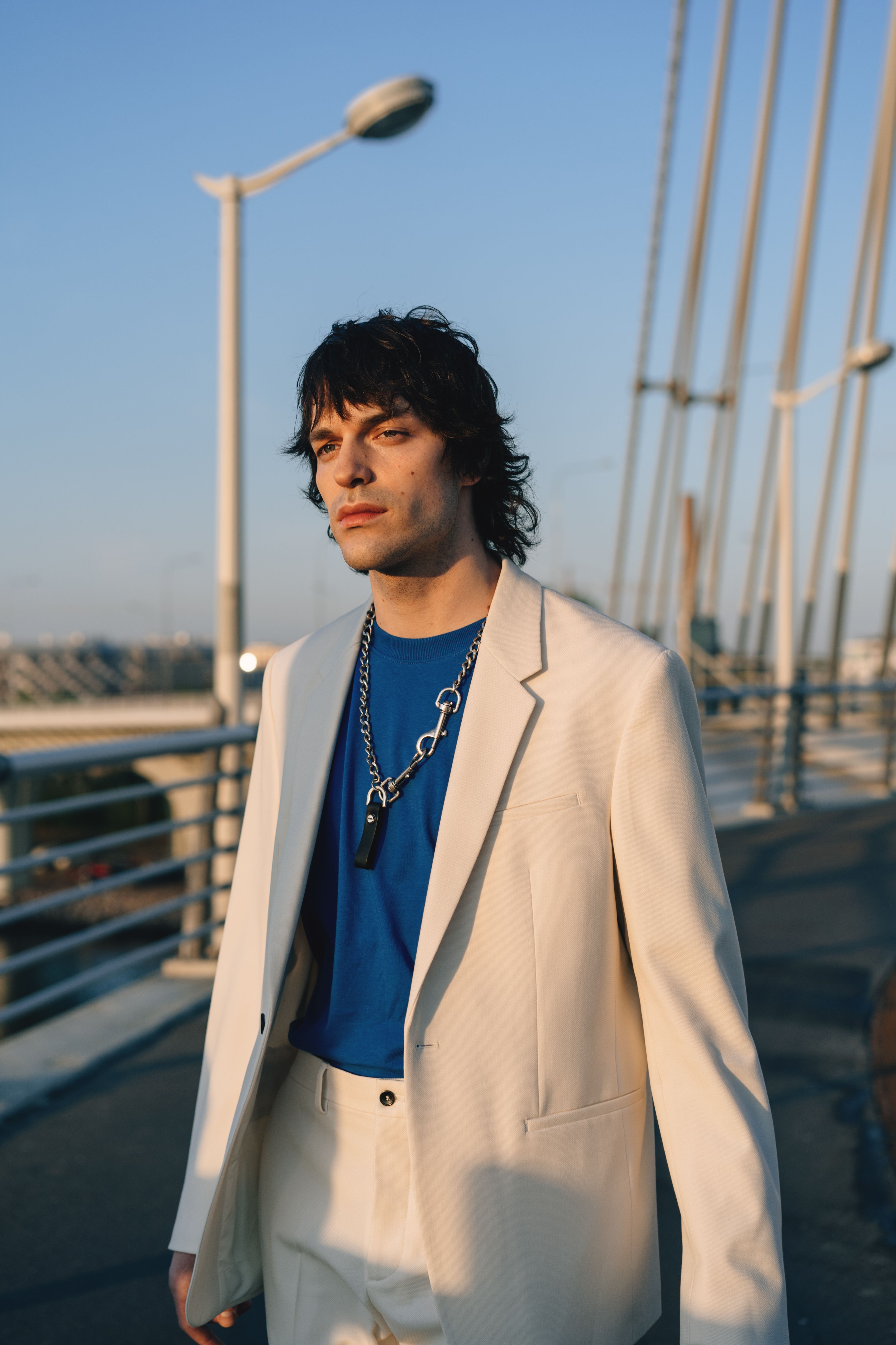 Stylish man in white suit and blue shirt walking on a city bridge during sunset.