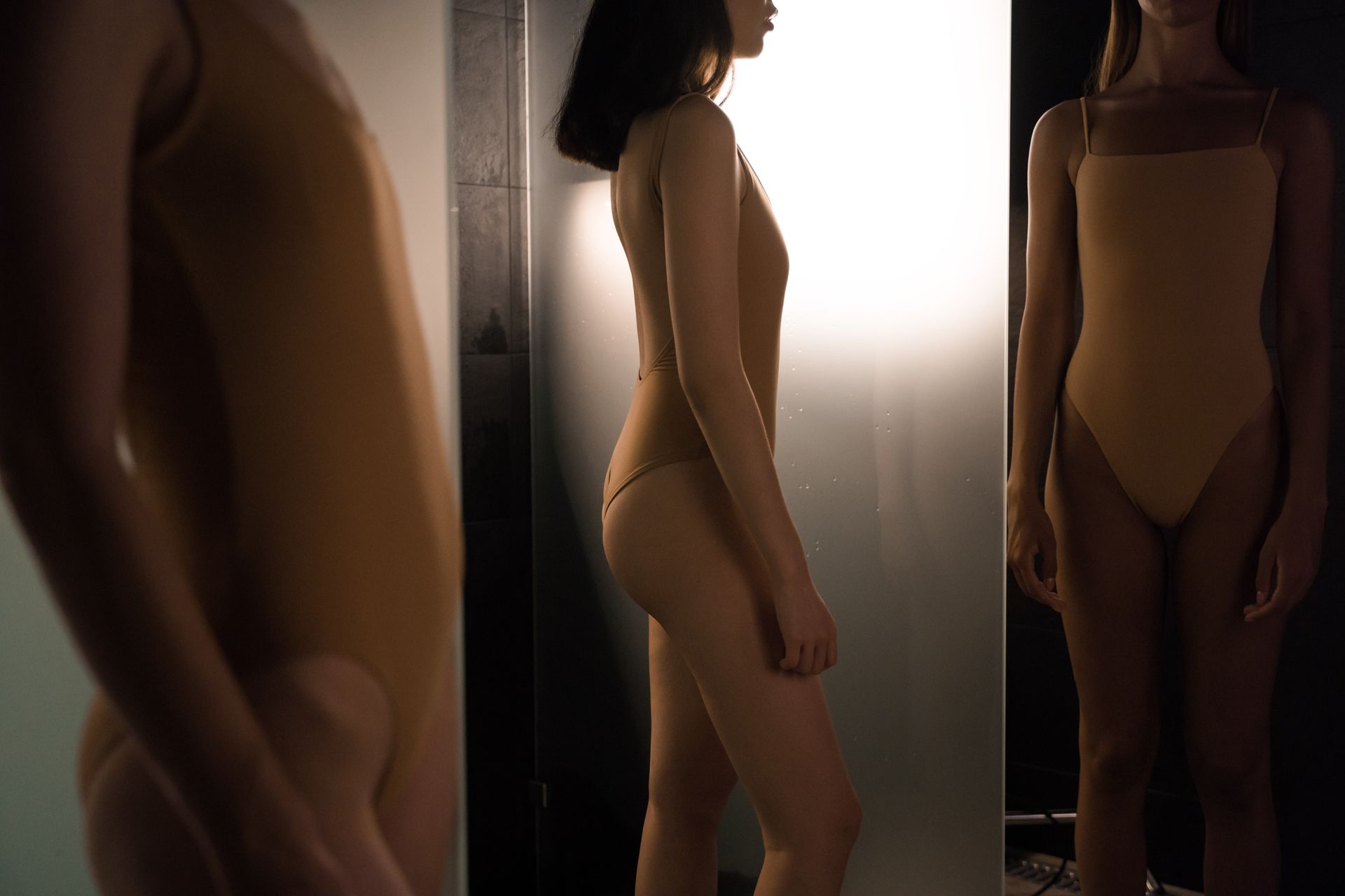 Two women in swimsuits stand near a glass panel with dramatic lighting.
