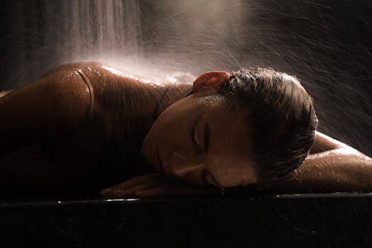 Woman in a spa setting under waterfall, relaxing with eyes closed on a marble surface.