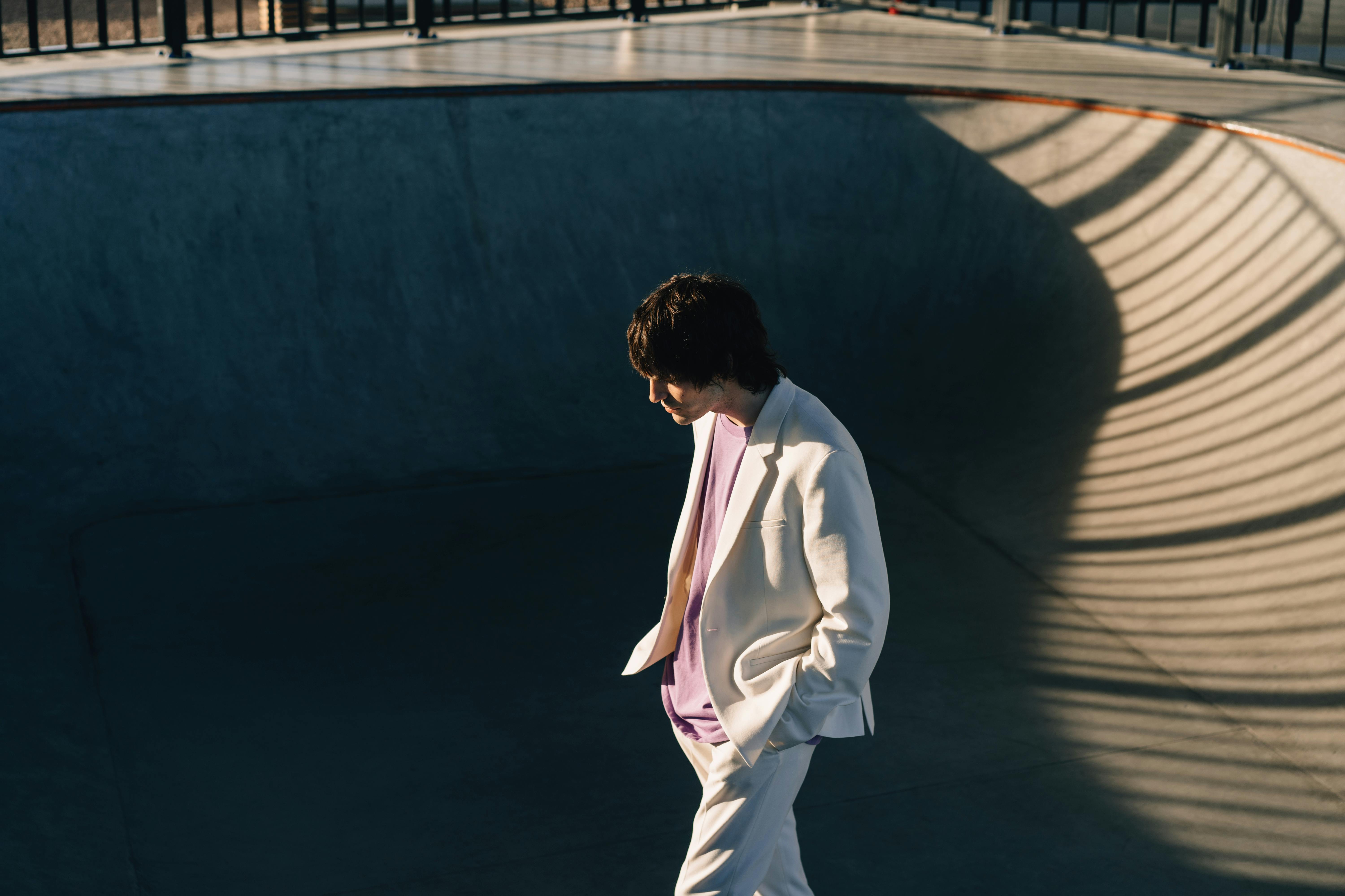 Fashionable man in white blazer walking in a sunlit skate park bowl.