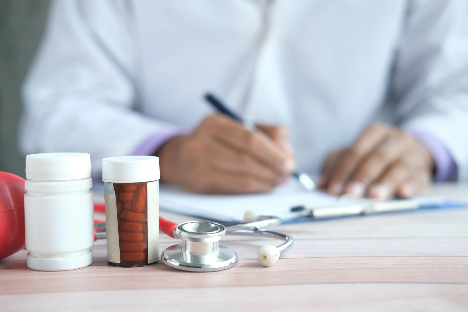 Close-up of doctor writing prescription with medicines and stethoscope on desk.