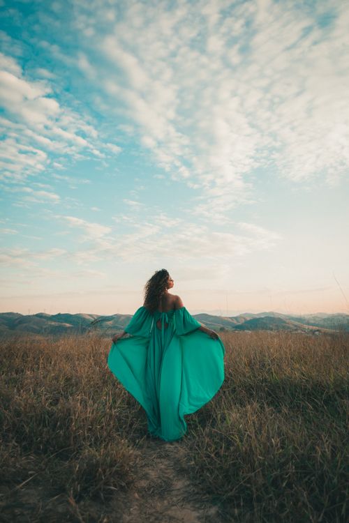 Free Woman in flowing green dress standing in a scenic grassy field against a blue sky in Volta Redonda, Brazil. Stock Photo