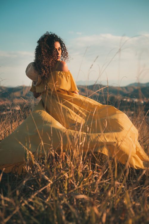 Free A woman in a flowing yellow dress stands in a field under a clear sky. Stock Photo