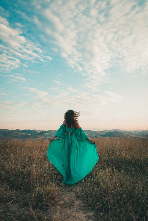 Free A woman in a flowing green dress stands on a grassy hill, embracing nature under a vast sky. Stock Photo