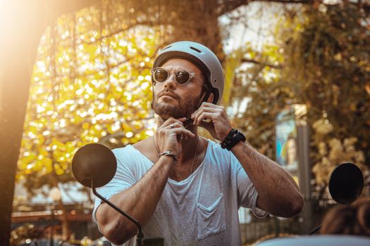 A man wearing sunglasses and a helmet adjusts his strap outdoors in bright sunlight.