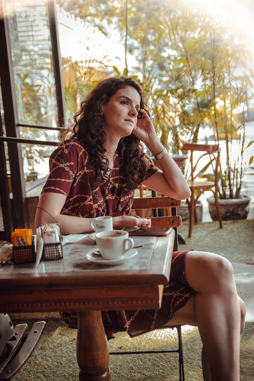Free A Woman in Printed Dress Sitting Near the Wooden Table with Her Legs Crossed Stock Photo