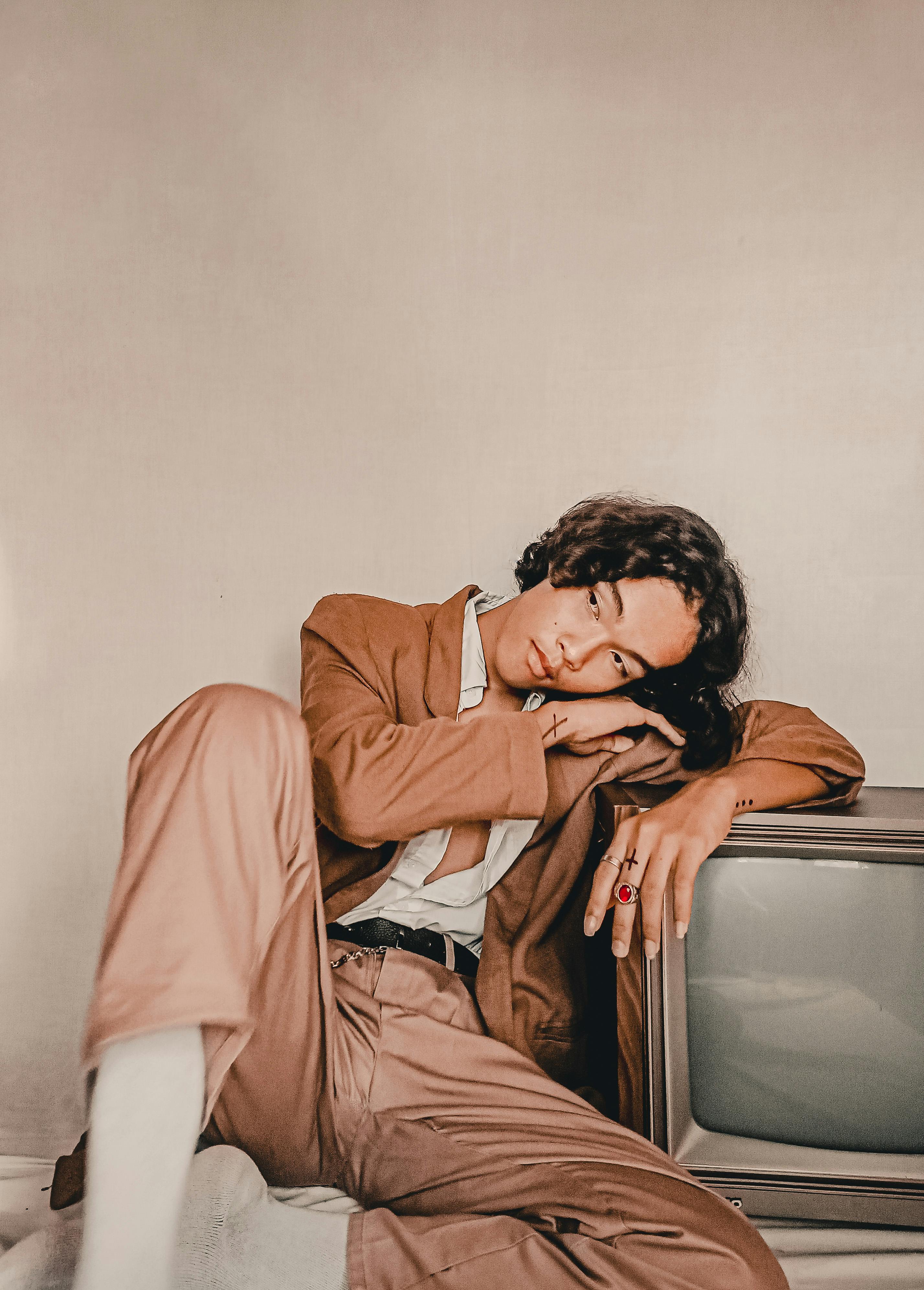 A stylish young adult in a brown outfit poses with a vintage TV on a minimalist backdrop.