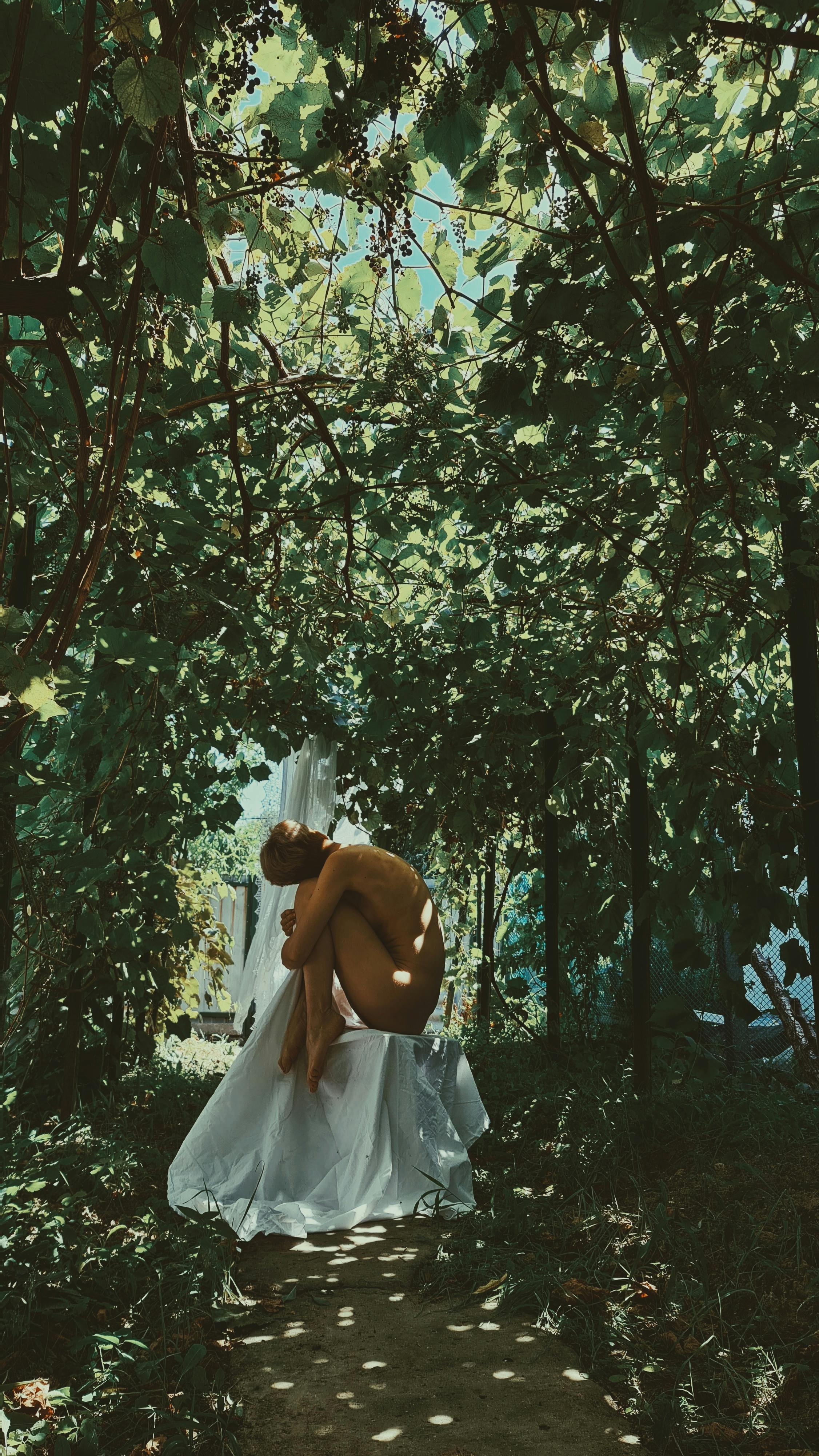 a naked person sitting on a white cloth near the green trees