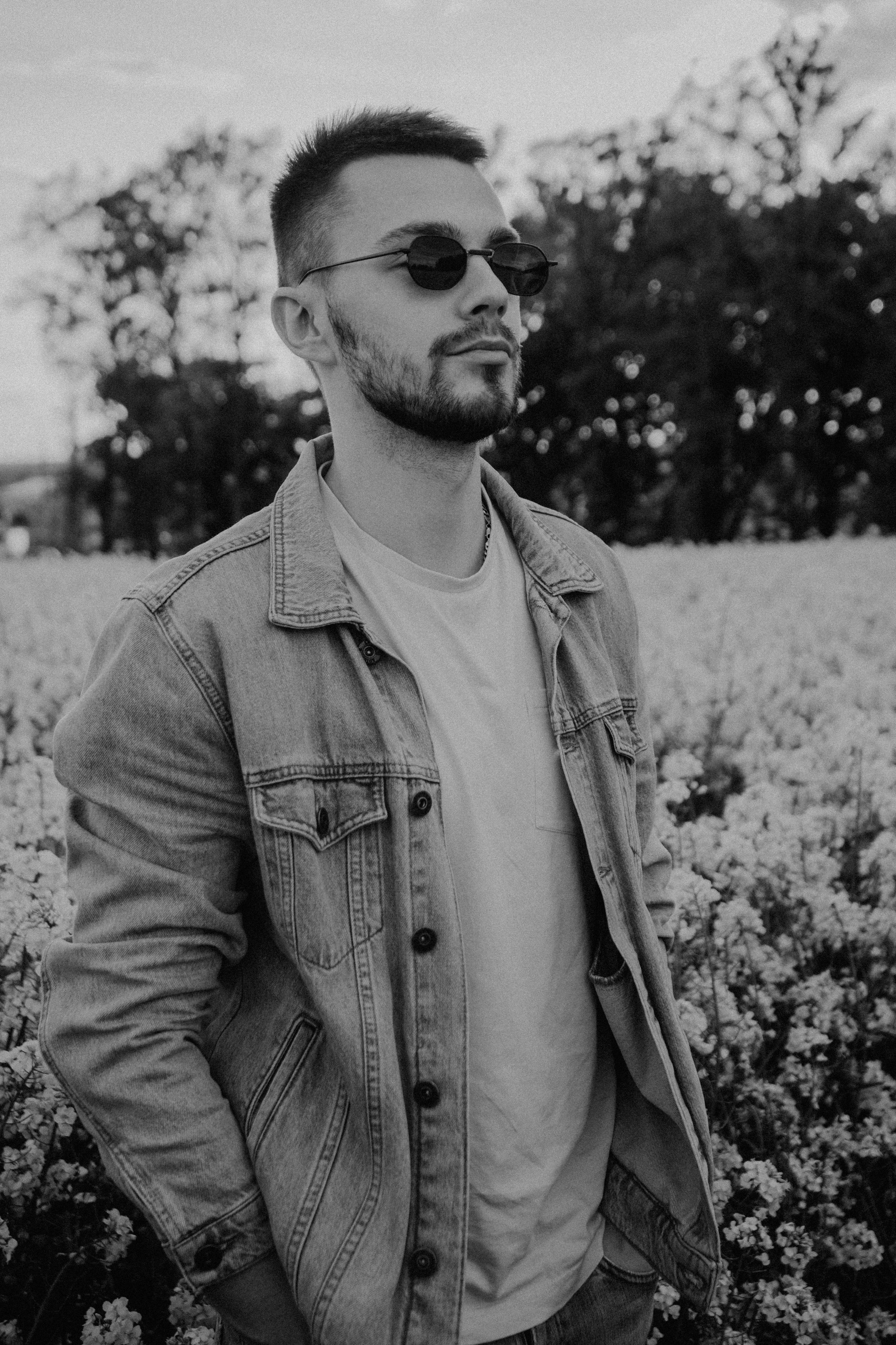 A bearded man wearing sunglasses and a denim jacket stands confidently in a black and white flower field, displaying modern style.