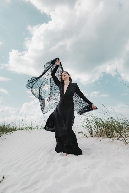 Free A woman in a black dress gracefully poses on New Smyrna Beach with a lace scarf against a cloudy sky. Stock Photo