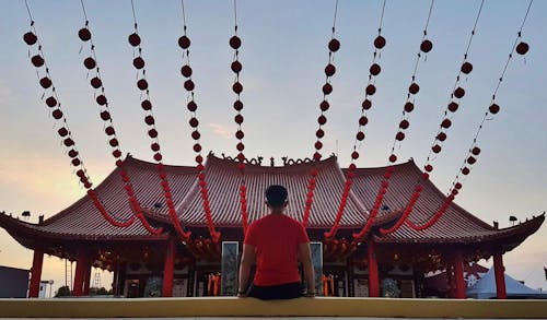 Man Sitting on Yellow Concrete Pavement Infront of Red Pagoda