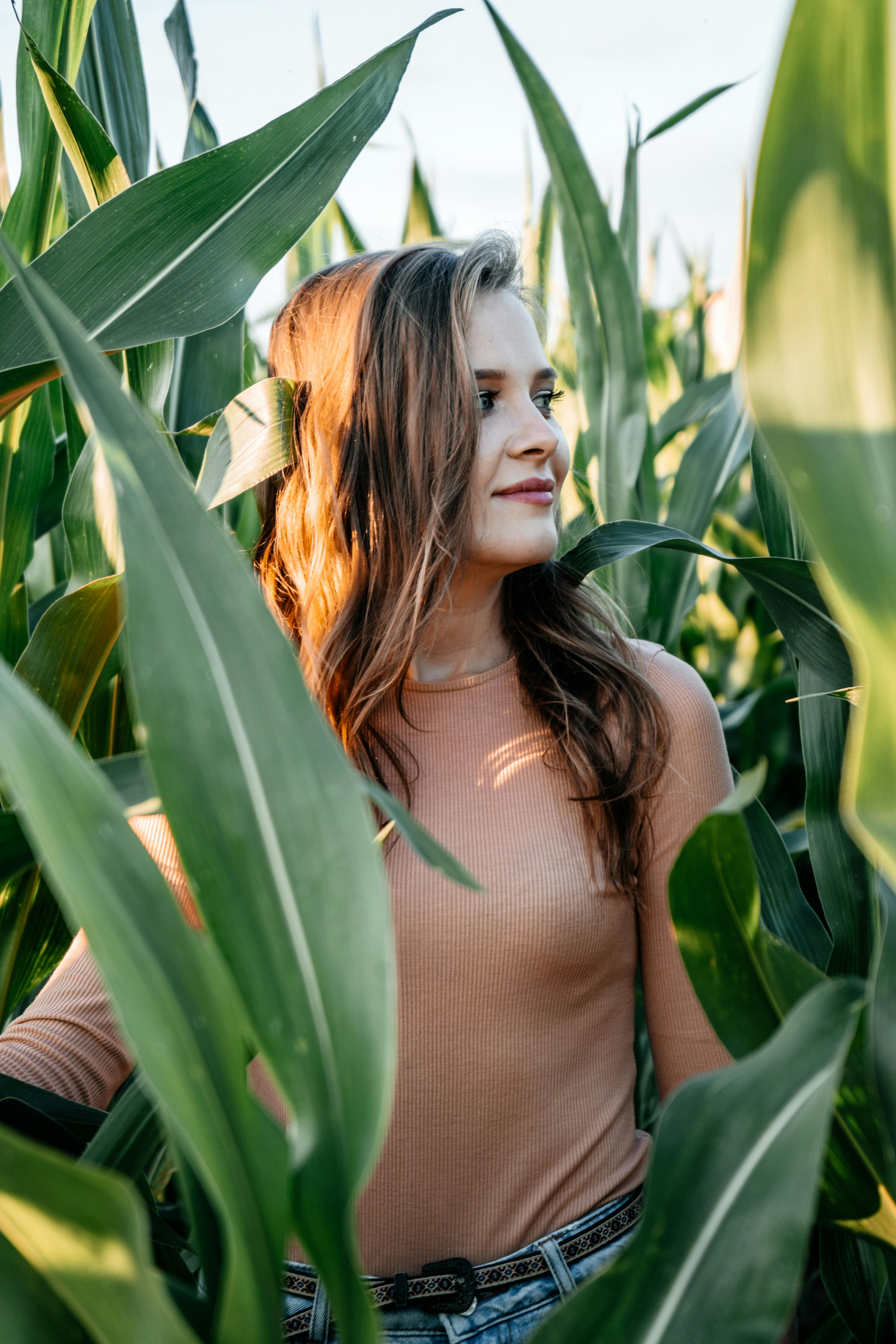 A young woman stands happily surrounded by tall corn plants in a rural field.