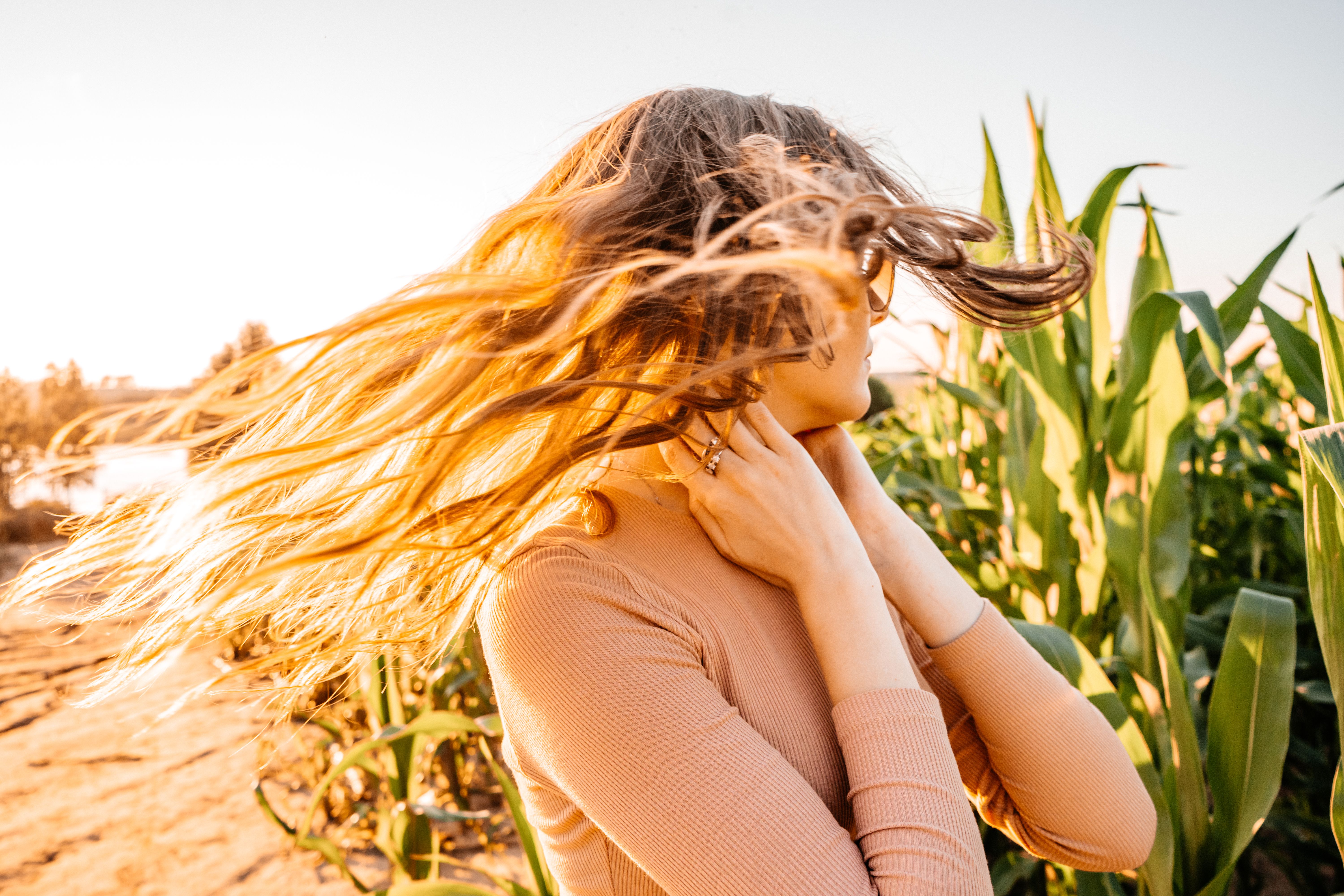 A woman with flowing hair stands in a cornfield during sunset, embracing nature.